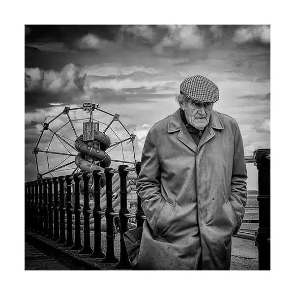 A black and white photo of an old man standing near a ferris wheel.