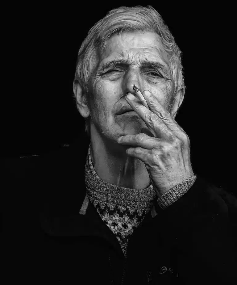 Faces_Photo_by John Gill-43 A black and white photo of an old man smoking a cigarette.