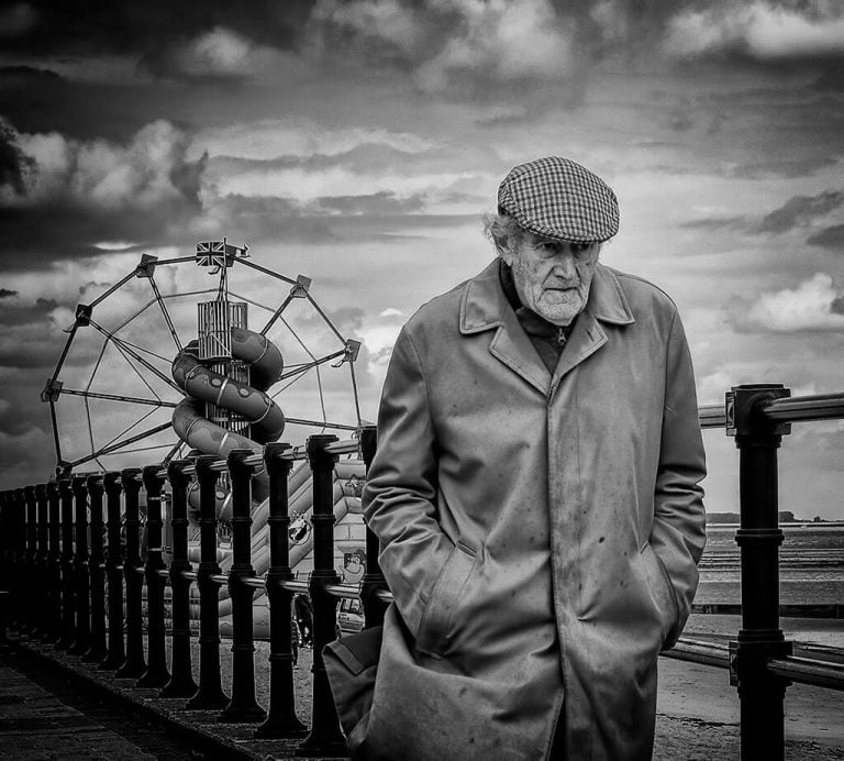 An old man standing on a railing near a ferris wheel.