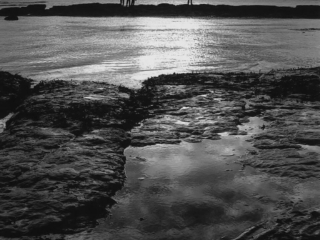 A black and white photo of people standing on rocks at the beach.