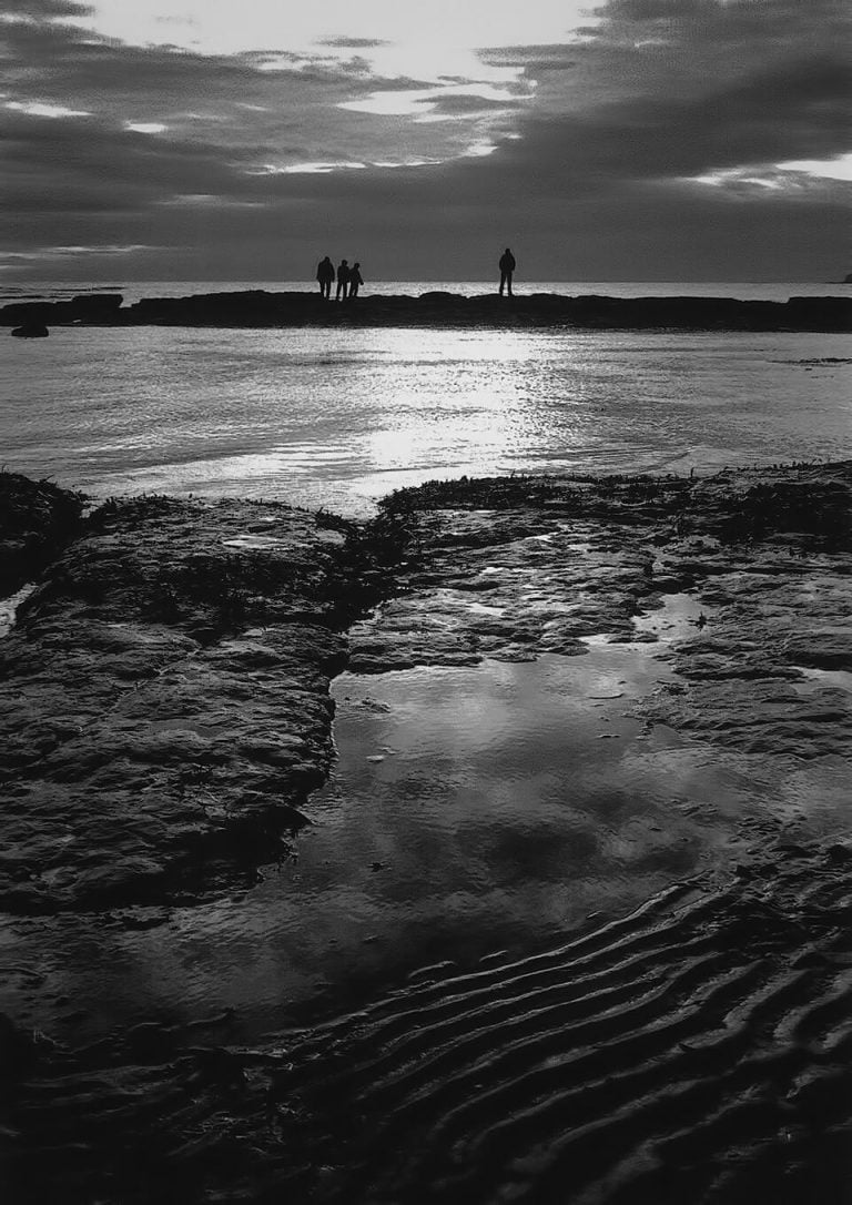 A black and white photo of people standing on rocks at the beach.