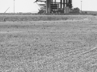 A man sitting on a beach in front of an oil refinery.