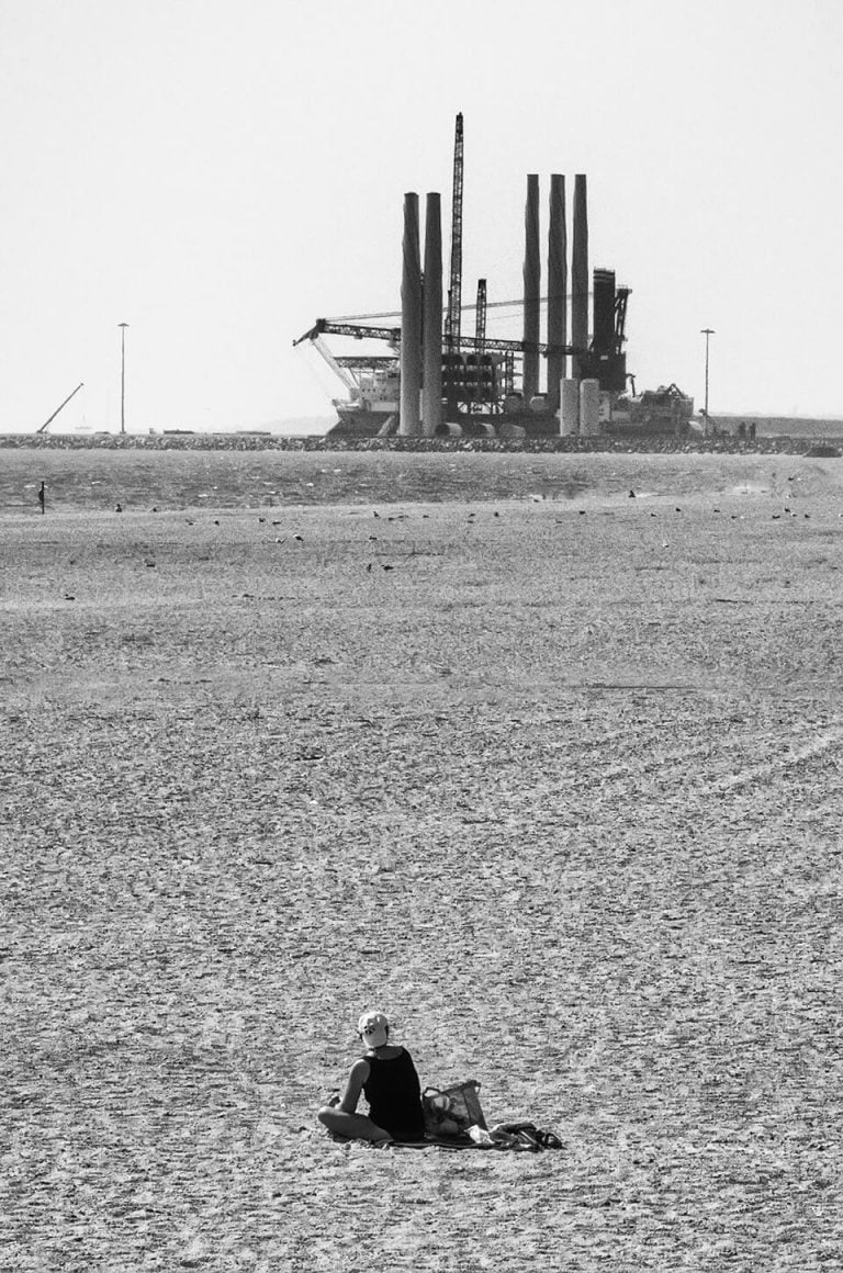 A man sitting on a beach in front of an oil refinery.