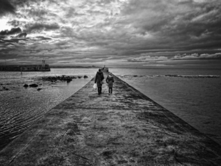 A black and white photo of two people walking on a pier. 