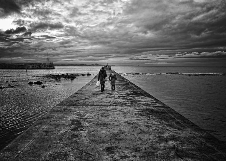 A black and white photo of two people walking on a pier. 