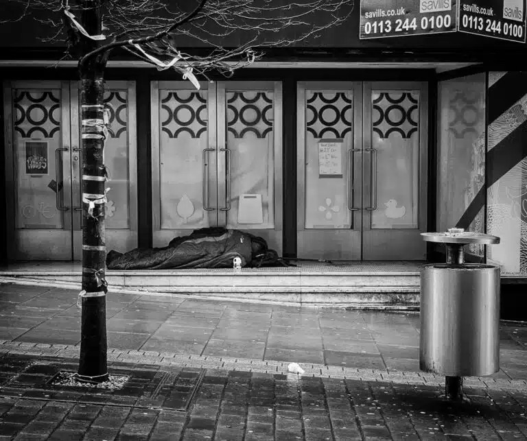 A black and white photo of a homeless man sleeping on a sidewalk.
