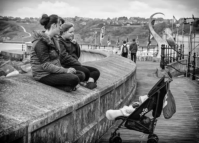 Two women sitting on a pier with a baby in a stroller.