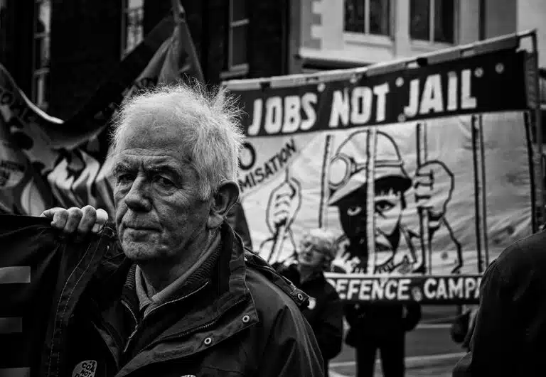 An old man holding a sign that says jobs not jail.