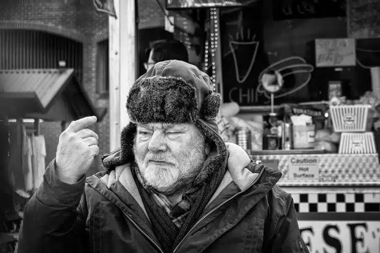 Street_Photography_Leeds_by John Gill-10 A man in a hat is standing in front of a food stand.