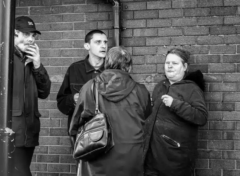 Street_Photography_Leeds_by John Gill-11 Black and white photograph of a group of people standing near a brick wall.