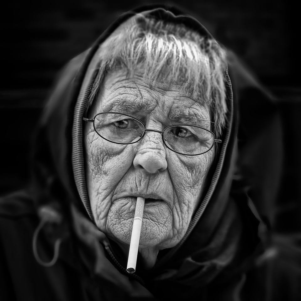 An elderly woman with glasses and a hood smokes a cigarette, looking off to the side. The image is in black and white.