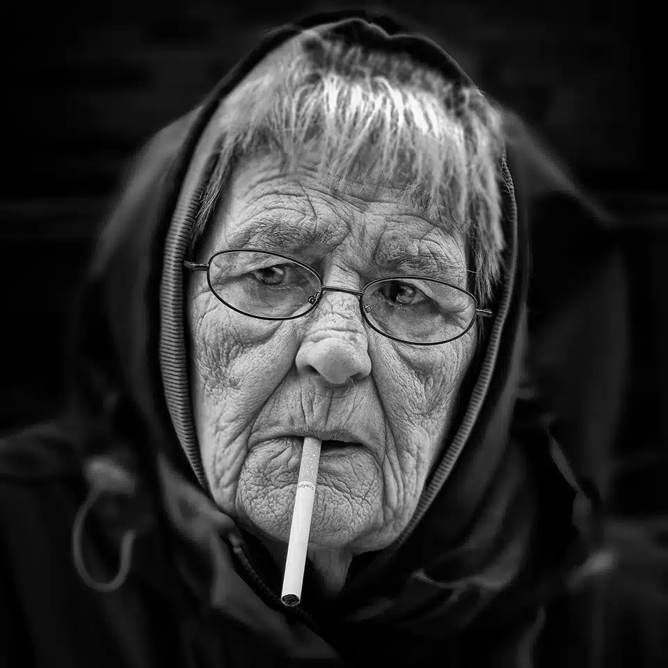 An elderly woman with glasses and a hood smokes a cigarette, looking off to the side. The image is in black and white.