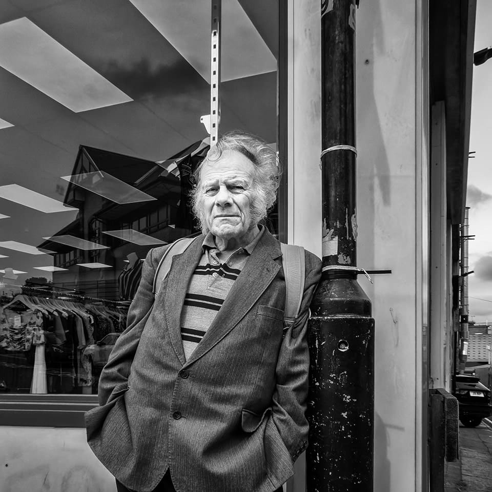 An older man in a suit and striped shirt leans against a pole outside a clothes shop, with buildings and clouds reflected in the glass behind him.