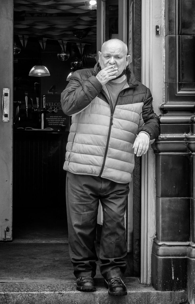 Black and white photograph of a man smoking outside a pub.