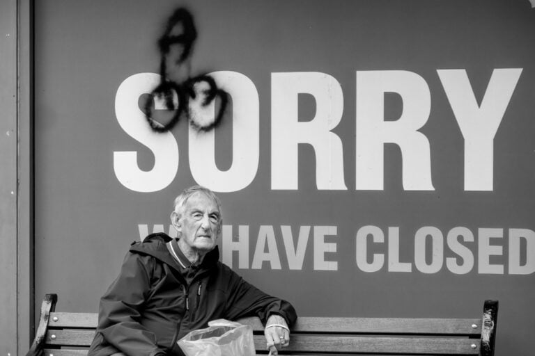 MAY A man sitting on a bench next to a sign that says sorry we have closed.