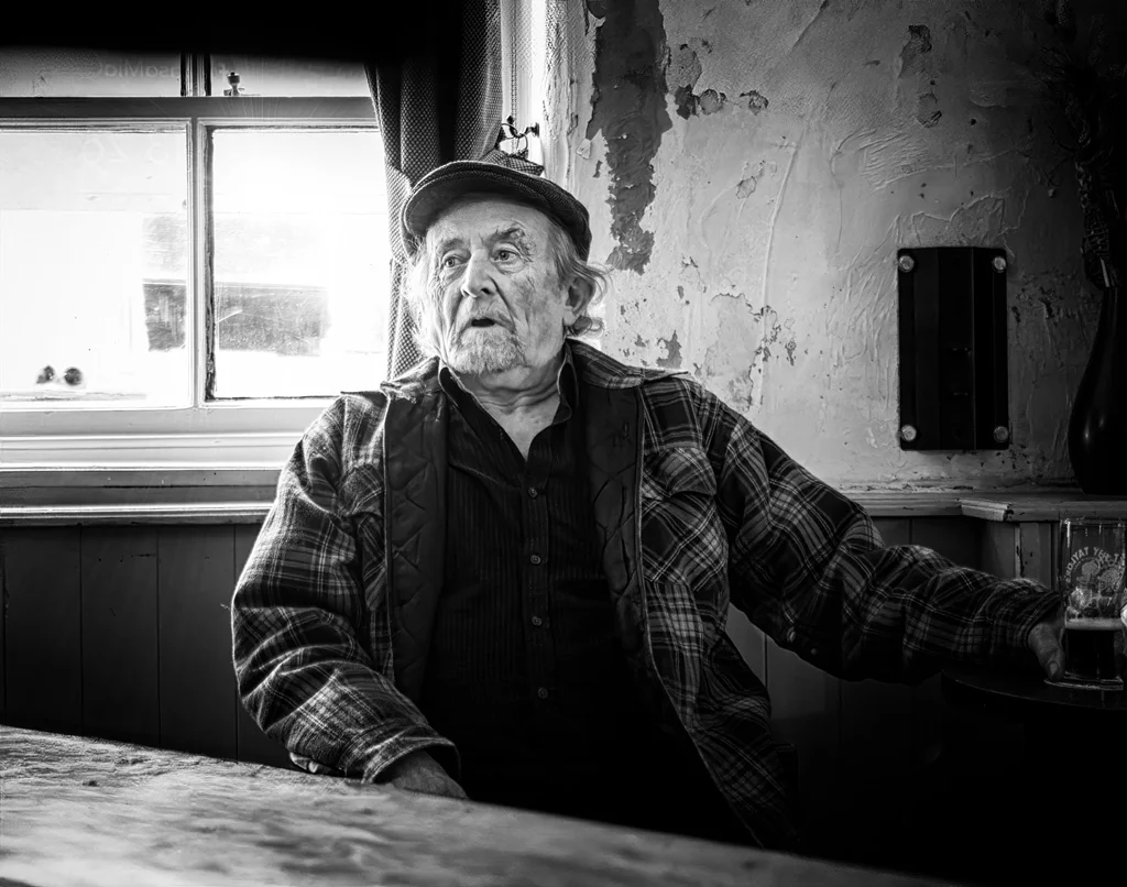 An old man sitting at a table with a beer.