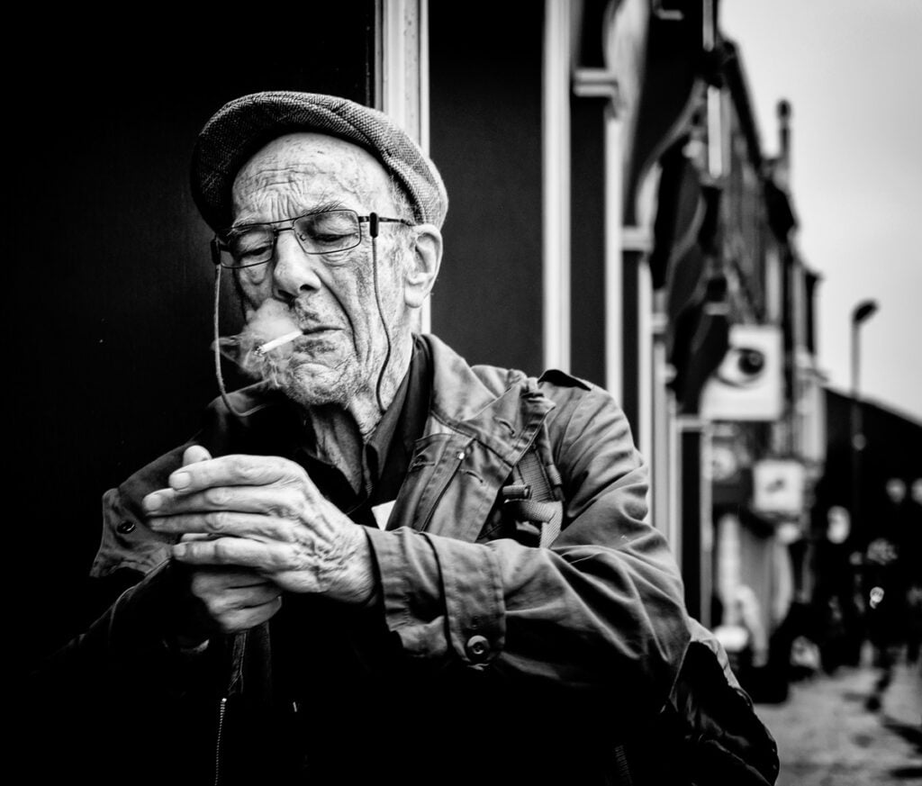 An old man smoking a cigarette in front of a building.