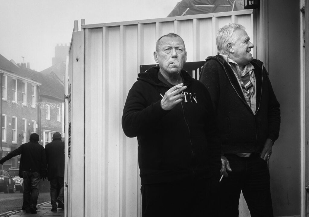 Two men standing in front of a building smoking a cigarette.