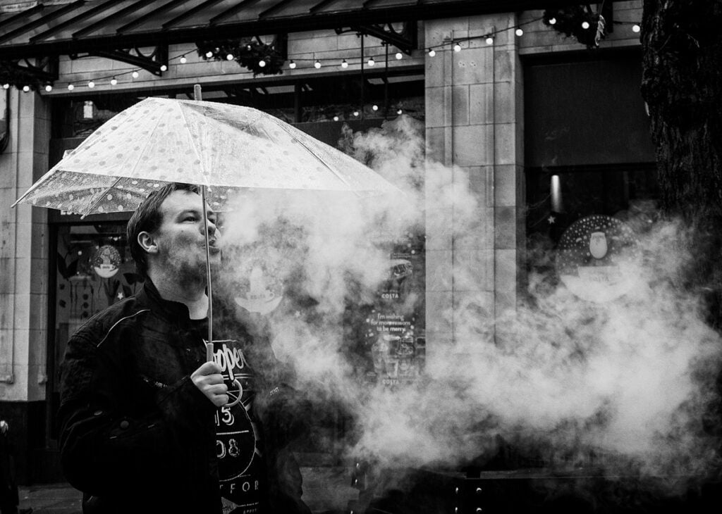A man smokes a cigarette under an umbrella.