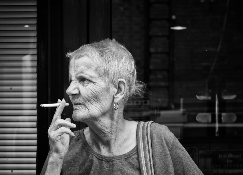 An old woman smoking a cigarette in front of a window.