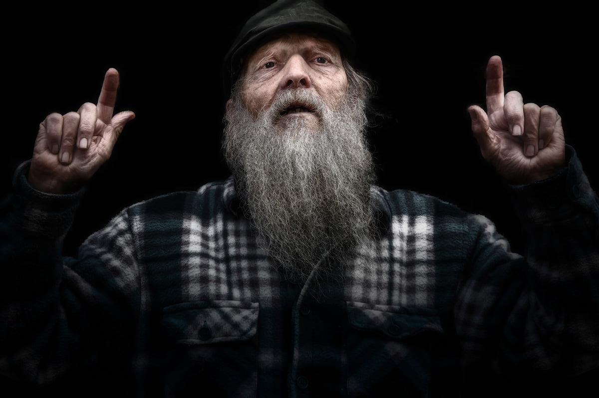 An elderly man with a long gray beard, wearing a plaid shirt and a dark cap, raises both index fingers against a black background.