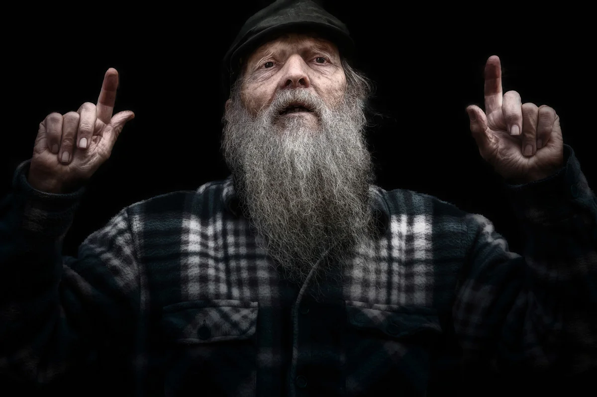It's not all black and white - some colour photography 3 An elderly man with a long gray beard, wearing a plaid shirt and a dark cap, raises both index fingers against a black background.