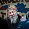 An elderly man with a gray beard wears a colorful knit hat and stands in front of a sign with partially visible text that includes “CANDLE” and “POP! POP!” while talking to unseen people. An elderly man with a gray beard wears a colorful knit hat and stands in front of a sign with partially visible text that includes "CANDLE" and "POP! POP!" while talking to unseen people.