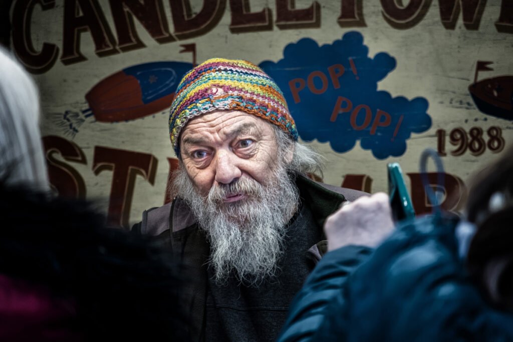 An elderly man with a gray beard wears a colorful knit hat and stands in front of a sign with partially visible text that includes "CANDLE" and "POP! POP!" while talking to unseen people.