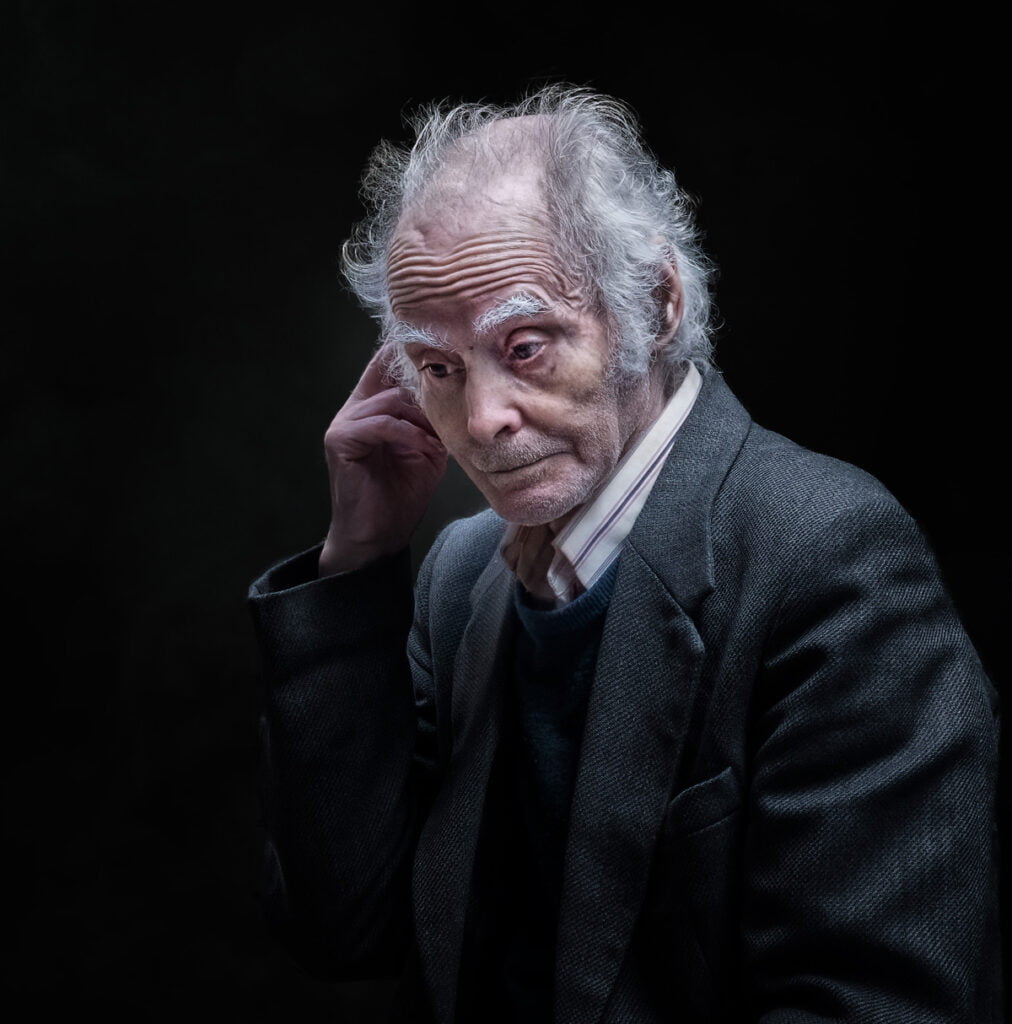 An elderly man with white hair, wearing a suit and sweater, touches his ear and looks down pensively against a dark background.