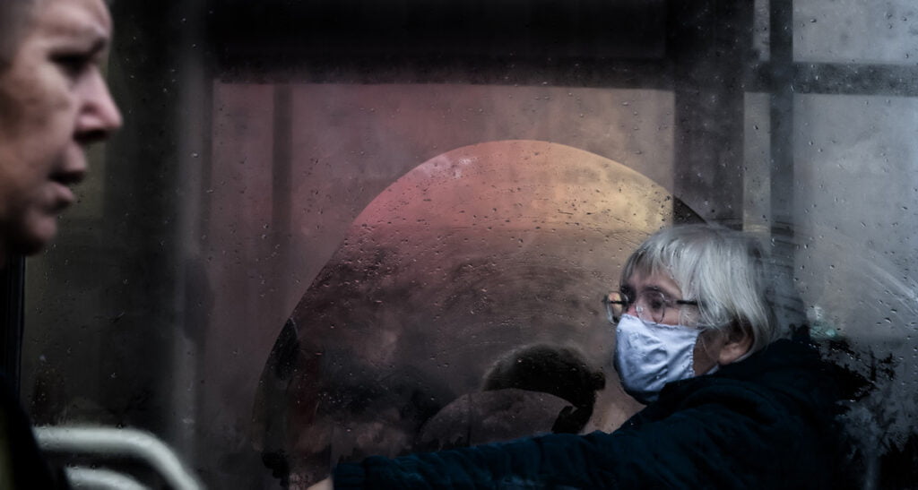 A person in a surgical mask is visible through a foggy window, while another person stands in the foreground. The scene appears to be inside a bus or train.