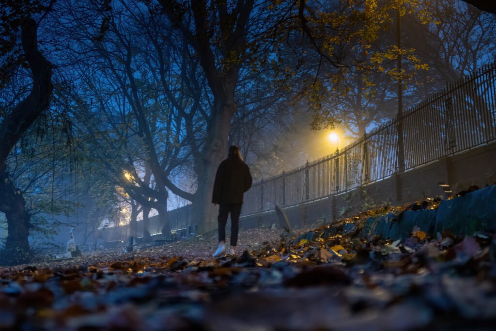 Person walking along a leaf-strewn path under trees at night, illuminated by streetlights with a foggy atmosphere.