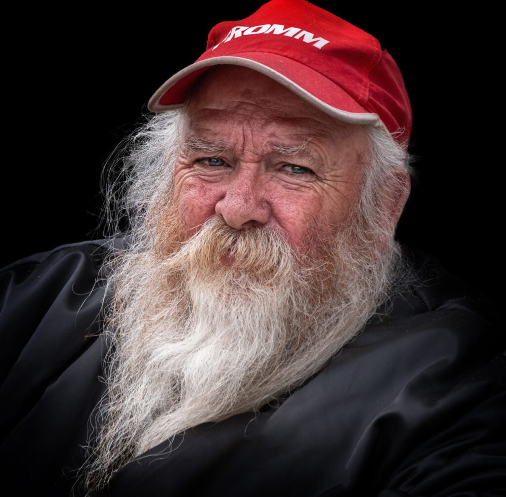 Elderly man with a long white beard wears a red cap and a black jacket. The background is black.