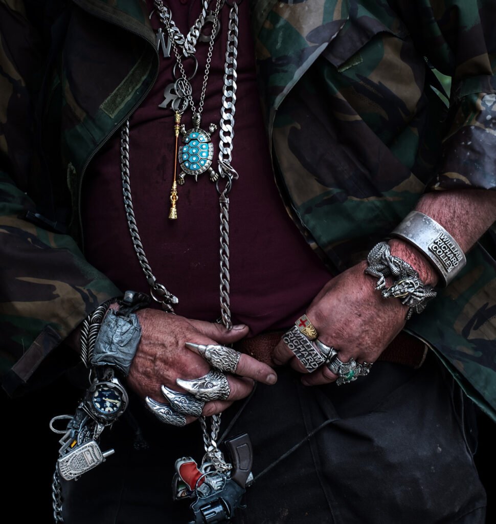A person wearing camo, a maroon shirt, and numerous large metal rings, bracelets, chains, and necklaces.