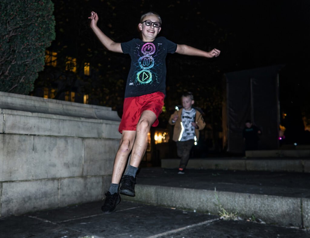 A boy wearing glasses, a black shirt, and red shorts jumps with arms outstretched at night while another child is visible in the background.