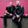 Two men in black leather outfits sit on a stone structure against a pink background, engaging in conversation. Two men in black leather outfits sit on a stone structure against a pink background, engaging in conversation.