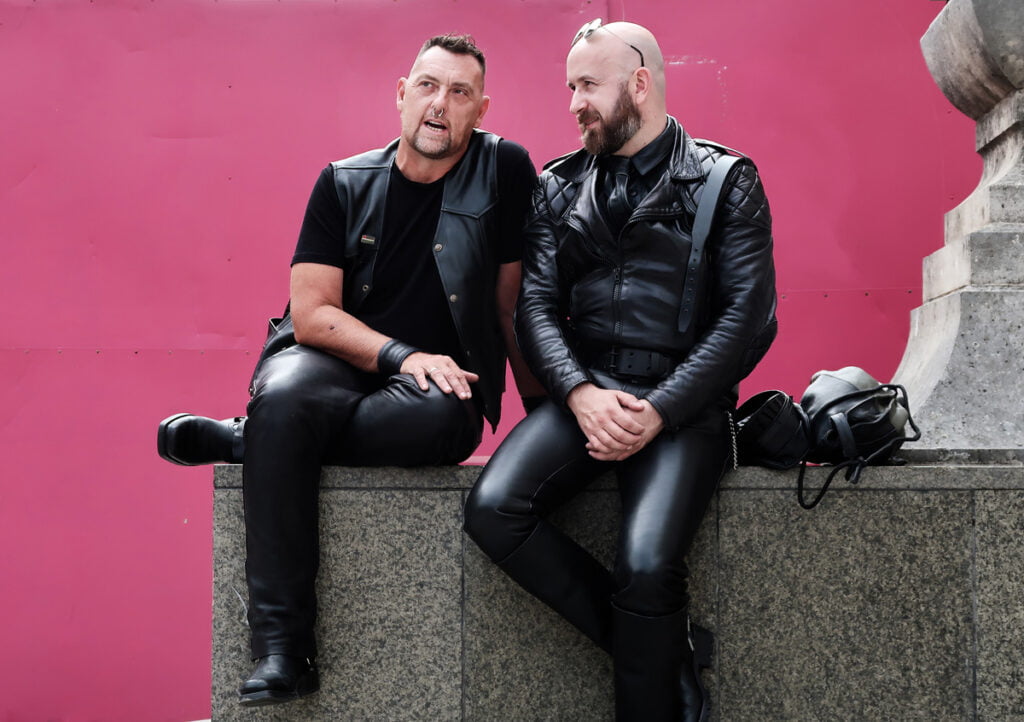 Two men in black leather outfits sit on a stone structure against a pink background, engaging in conversation.