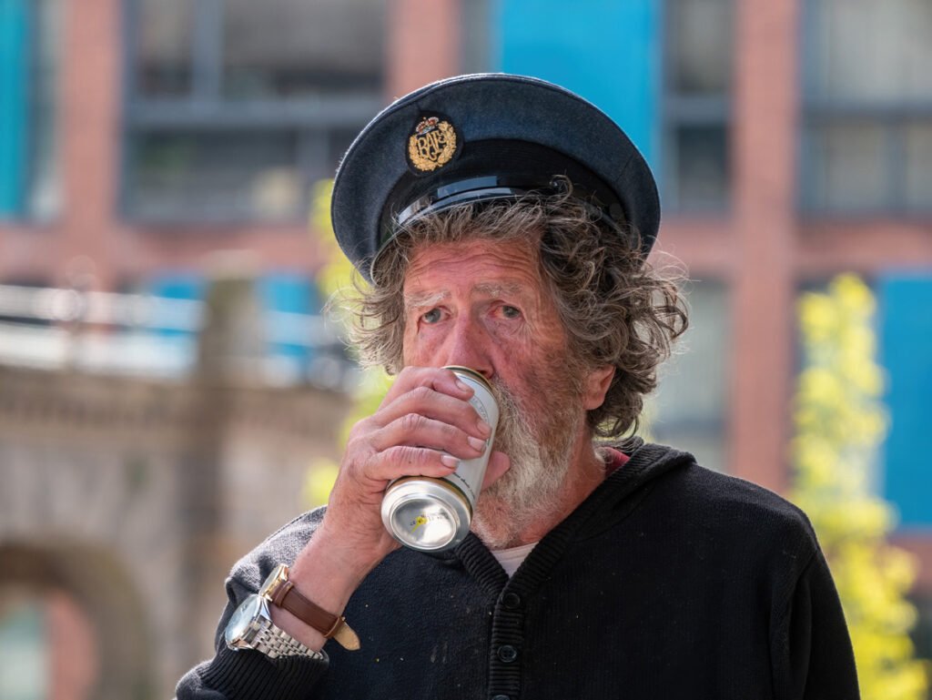 A man with curly hair and a beard wearing a dark hat and black sweater drinks from a can in an urban setting.