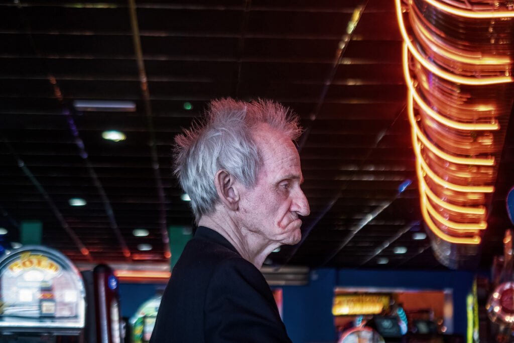An elderly person with white hair stands in profile inside a dimly lit space with bright neon lights and arcade games in the background.