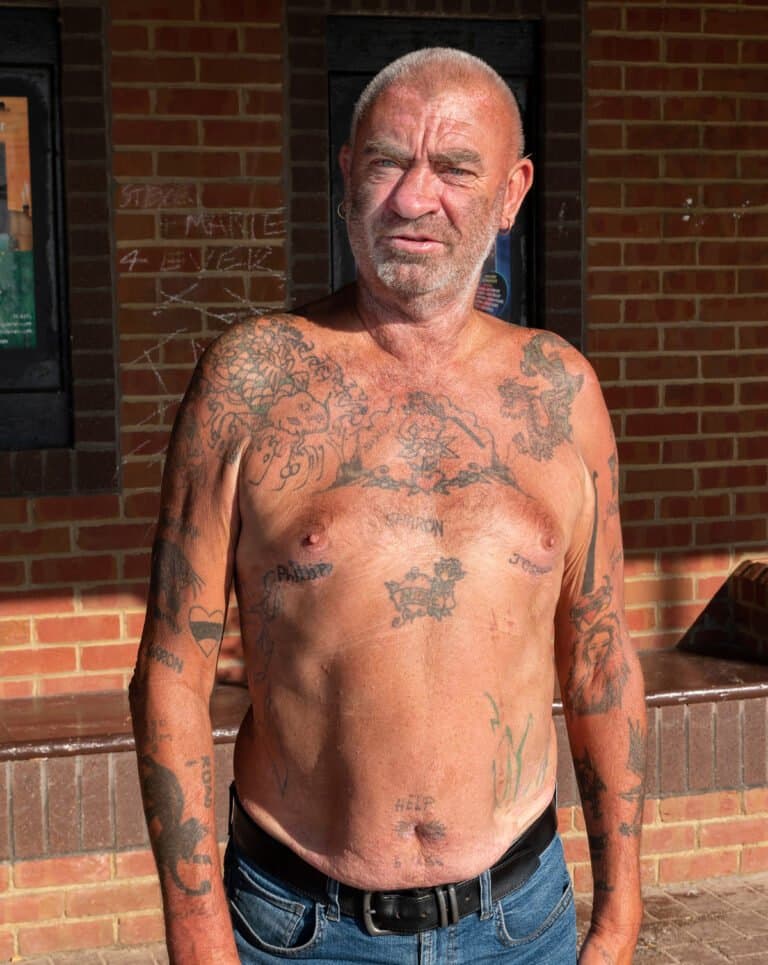 A shirtless man with numerous tattoos stands against a brick wall.