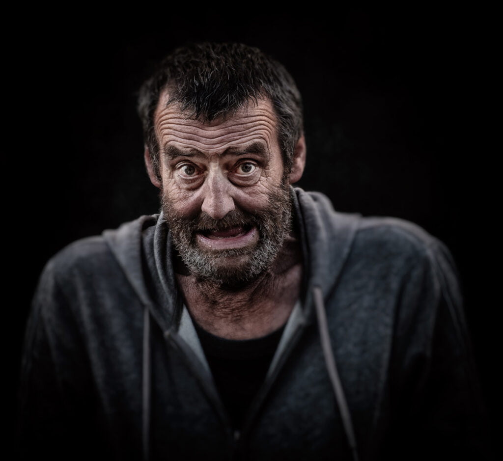 A man with a scruffy beard and a worn gray hoodie makes a grimacing facial expression against a dark background.