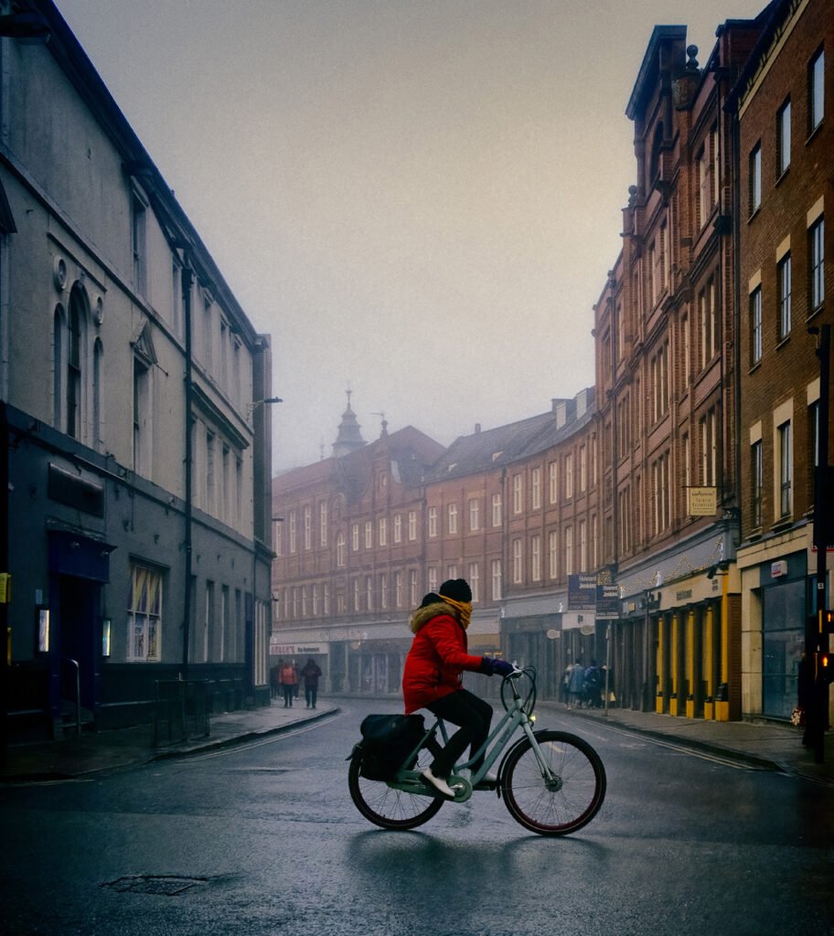 A person wearing a red jacket and yellow scarf rides a bicycle on a foggy street flanked by old brick buildings.