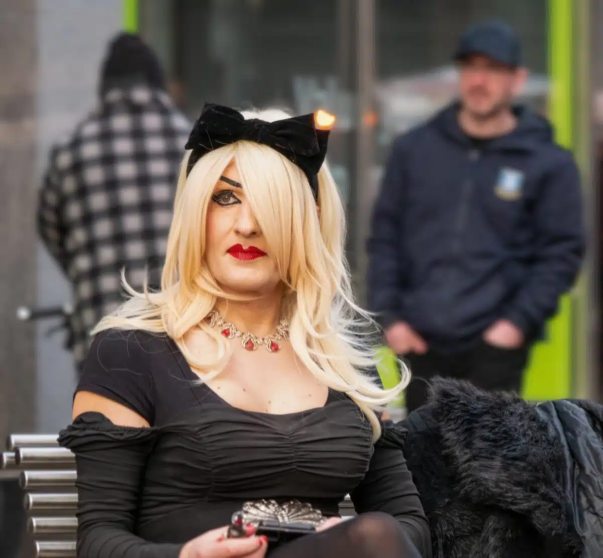 A person with long blonde hair, wearing a black dress, black bow headband, and red necklace, sits on a bench holding a small clutch. Two other people are blurred in the background.