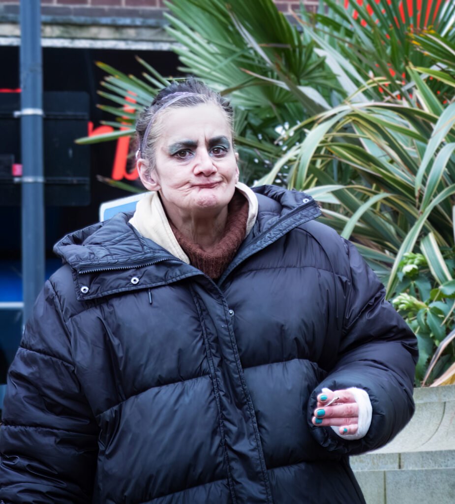 A woman with heavy makeup and a black puffer jacket stands outdoors beside green plants, holding a cigarette.