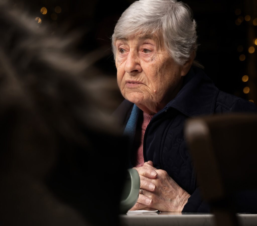 An elderly woman with short gray hair sits at a table, resting her hands on a green mug, looking slightly to the side with a contemplative expression. She wears a dark jacket indoors.