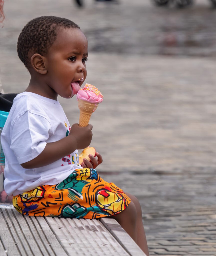 A child in colorful shorts and a white shirt sits on a stone bench, enjoying a pink ice cream cone with sprinkles.