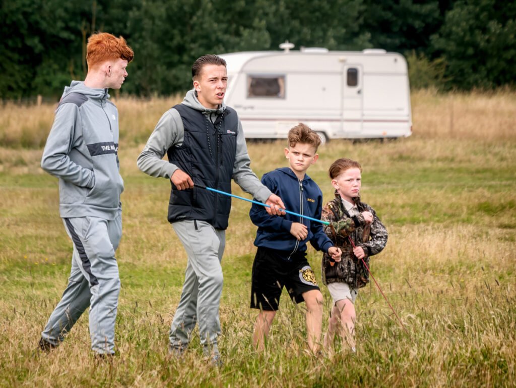 Four boys walk through a grassy field, with three of them holding leashes. A white camper van is visible in the background.