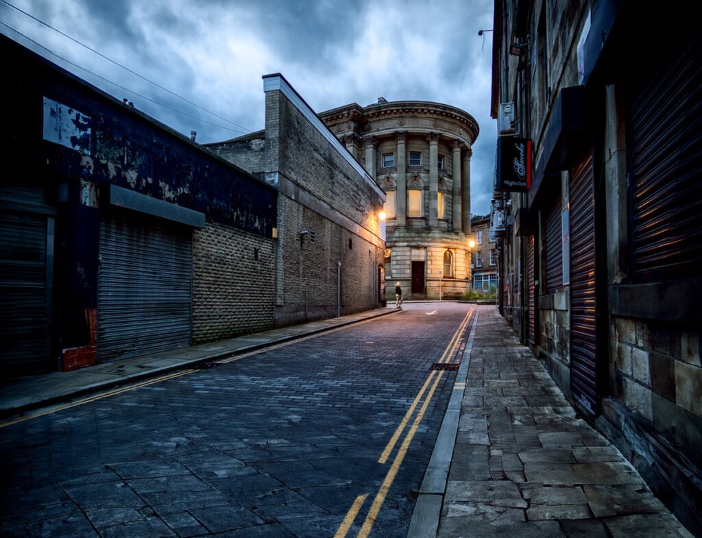 A narrow, empty cobblestone street flanked by old buildings on both sides leads to a round, illuminated structure under a moody, cloudy sky. The street is lined with closed shop shutters.