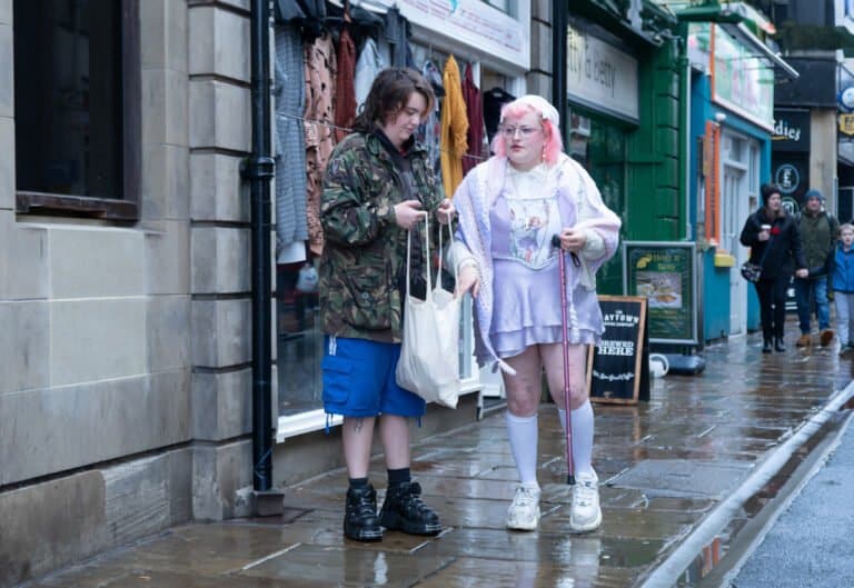 Two people stand on a wet sidewalk in front of a shop. One wears a camouflage jacket and checks out a bag, while the other, in pastel attire, holds a leash without a visible pet.