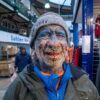 A person with numerous facial piercings, tattoos, and body modifications stands in a market, smiling and wearing a York beanie, blue shirt, and green jacket. A person with numerous facial piercings, tattoos, and body modifications stands in a market, smiling and wearing a York beanie, blue shirt, and green jacket.