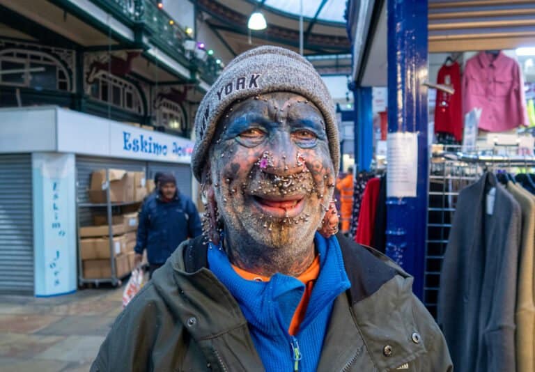 A person with numerous facial piercings, tattoos, and body modifications stands in a market, smiling and wearing a York beanie, blue shirt, and green jacket.